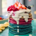 Stack of colorful ombre pancakes in blue, pink, and purple hues served on a plate with fresh fruit and syrup.
