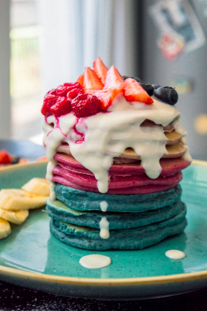 Stack of colorful ombre pancakes in blue, pink, and purple hues served on a plate with fresh fruit and syrup.