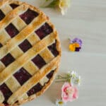 Homemade vegan cherry pie with golden lattice crust, decorated with edible flowers on a light wooden background