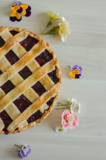 Homemade vegan cherry pie with golden lattice crust, decorated with edible flowers on a light wooden background