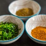 Small glass bowl of garam masala spice blend on a white kitchen counter with whole spices scattered nearby.