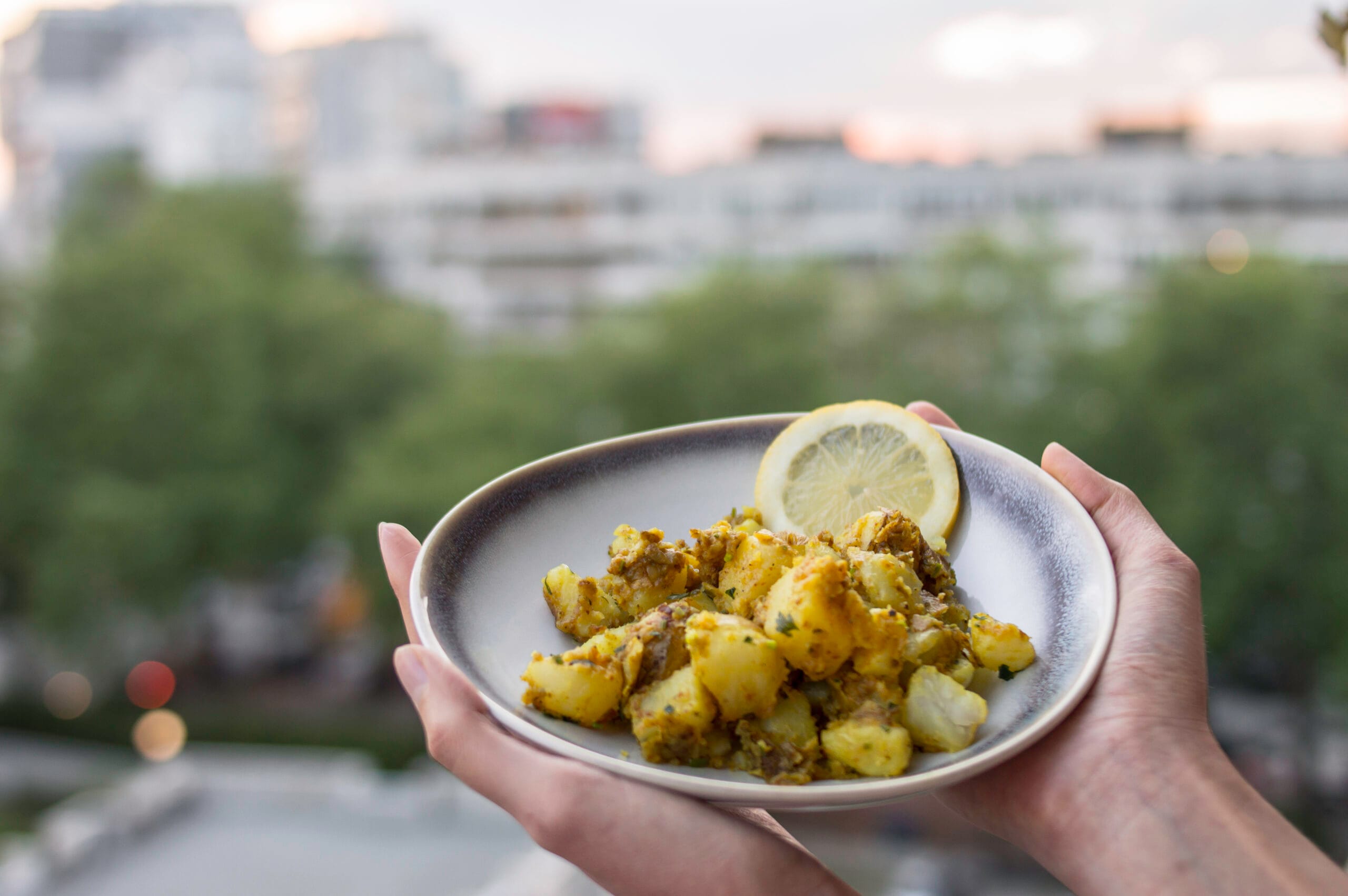 A bowl of golden Bombay potatoes served with lemon slices on a balcony table with a blurred cityscape in the background.