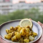 A bowl of golden Bombay potatoes served with lemon slices on a balcony table with a blurred cityscape in the background.