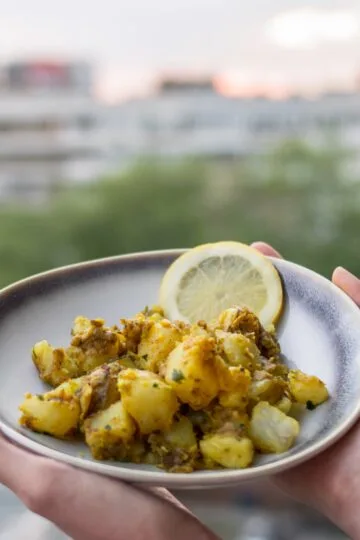 A bowl of golden Bombay potatoes served with lemon slices on a balcony table with a blurred cityscape in the background.