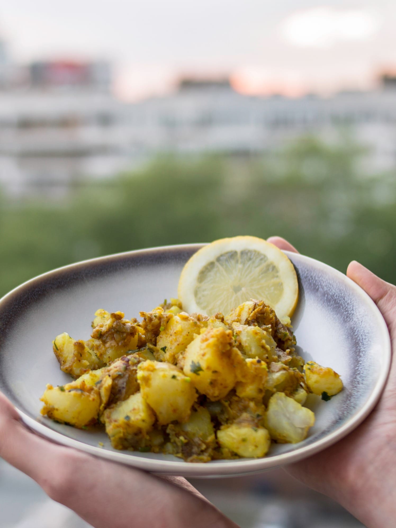 A bowl of golden Bombay potatoes served with lemon slices on a balcony table with a blurred cityscape in the background.