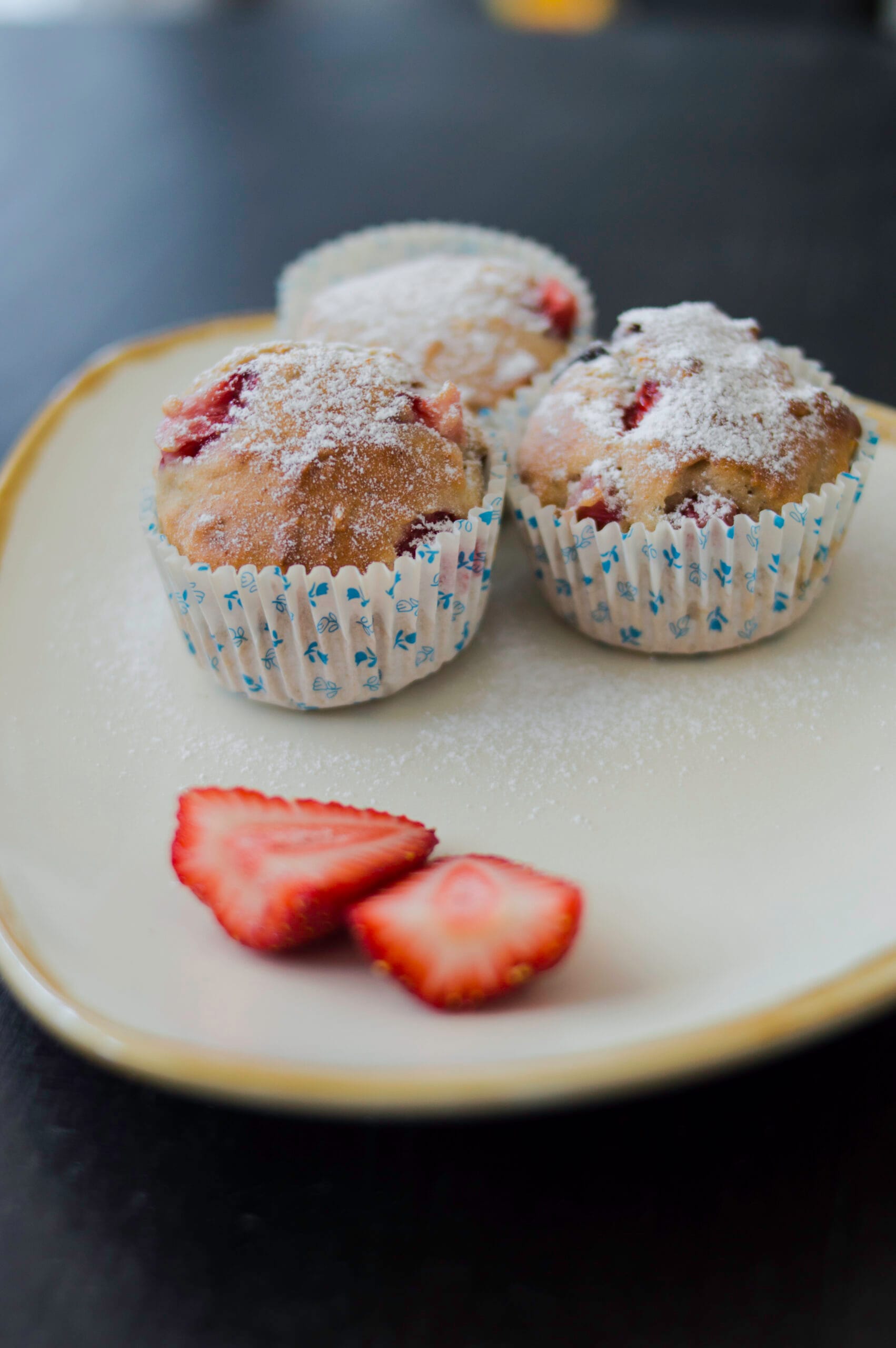 A batch of golden strawberry-banana muffins with melted chocolate chips on top, served with sliced strawberries on the side.
